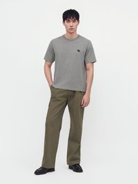 Homme portant un t-shirt gris à manches courtes avec un petit logo noir et un pantalon large vert olive avec des chaussures noires.