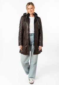 Manteau en cuir marron avec une capuche, doté d'une fermeture éclair frontale, de cordons élastiques et de deux poches latérales. Porté sur une chemise blanche et un jean clair.