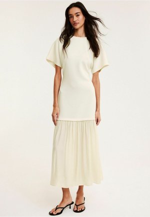 Kobieta stojąca w kremowej sukience midi z falbaniastymi rękawami i marszczoną spódnicą, ubraną w czarne sandały na niskim obcasie na białym tle.