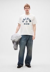 Valge puuvillane t-särk, millel on tumesinise tekstiga trükk "CALVIN KLEIN." Kantud helesiniste teksadega, hoides halli jakki ja musti kingi.