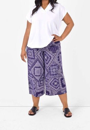 Vrouw staat met hand op heup, draagt een wit T-shirt met korte mouwen en V-hals, paarse wijde cropped broek met patroon en zwarte slippers.
