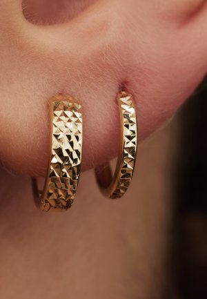 Boucles d'oreilles en forme de créoles en or avec un design texturé et facetté, et une finition polie, présentées sur une oreille. Les boucles d'oreilles affichent des formes symétriques et brillent.