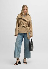 Tan double-breasted jacket with belt, light denim wide-leg jeans, and black strappy heels. Black handbag and long straight hair.