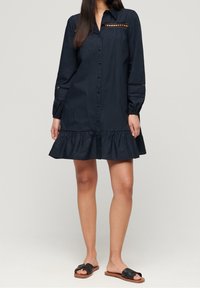 Navy Hemdkleid mit Rüsche, featuring lange Ärmel, Ausschnittdetails und einen strukturierten Schnitt. Kombiniert mit schwarzen flachen Sandalen.