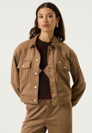 Frau steht mit den Händen hinter dem Rücken, trägt eine beigefarbene Jacke und Hose, einen dunklen Pullover mit beigem Rautenmuster und goldene Creolen.
