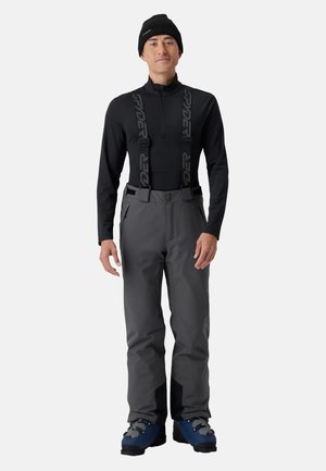Homme portant un haut noir à manches longues, un pantalon de ski gris avec des bretelles noires, des chaussures de ski bleues et un bonnet noir, debout devant un fond blanc.