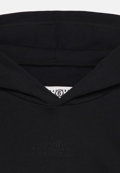 Κοντινή λήψη μαύρου φούτερ με κουκούλα που δείχνει την κουκούλα, μια λευκή ετικέτα μεγέθους με τον αριθμό 6 και ανάγλυφους αριθμούς διατεταγμένους σε τρεις σειρές στο στήθος.