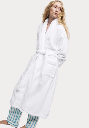 White terry cloth bathrobe with a shawl collar, tie belt, and front pockets; worn over light blue and white striped pants.