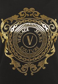 Černá látka se zlatým zdobeným kruhovým emblémem obsahujícím nápis "VERSACE JEANS COUTURE" a velkým písmenem "V" uprostřed.