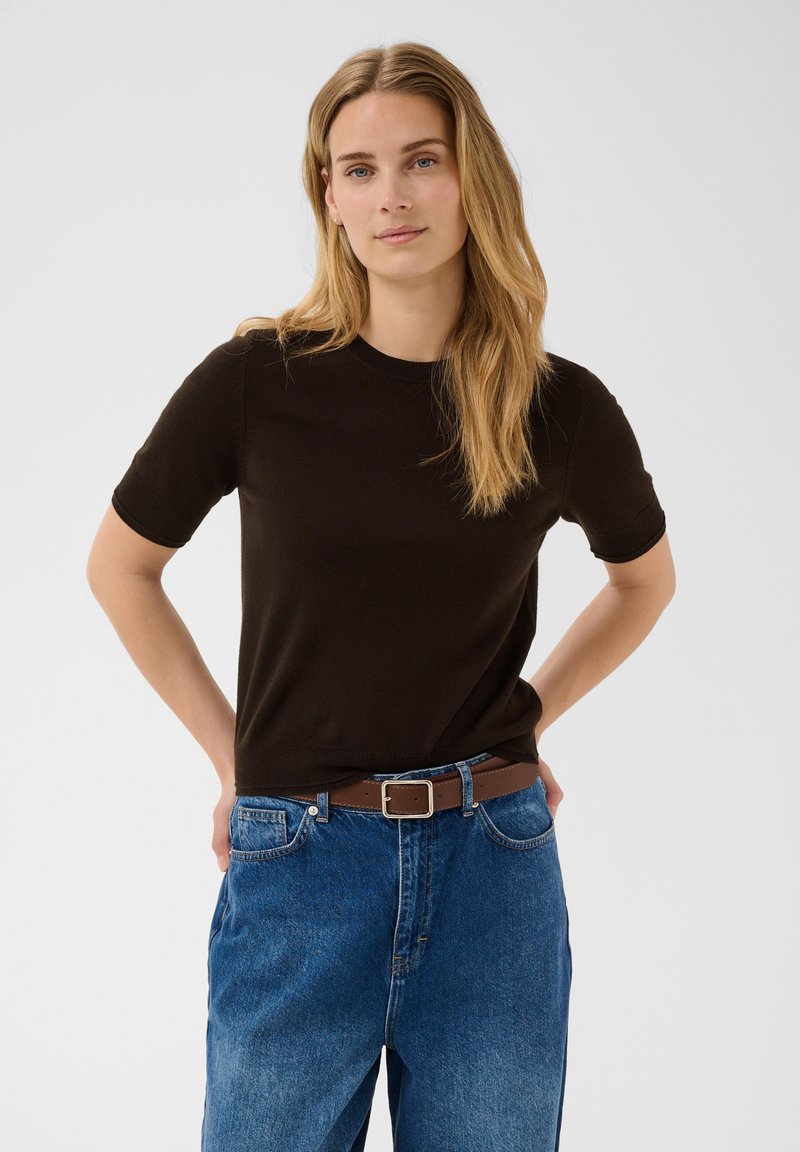 Femme aux longs cheveux blonds portant un haut noir à manches courtes rentré dans un jean bleu avec une ceinture marron, debout les mains derrière le dos.
