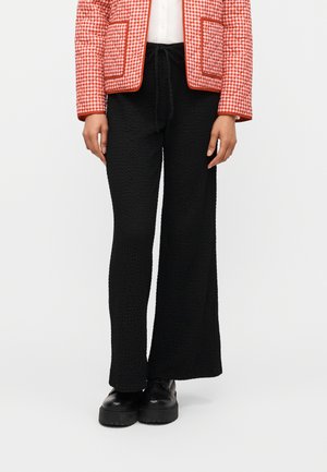 Person trägt schwarze, strukturierte, weit geschnittene Hose, schwarze klobige Schuhe, weißes Oberteil und rot-weiß karierten Blazer mit Taschen.