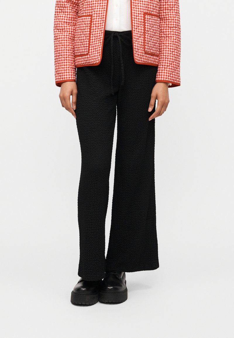 Άτομο που φοράει μαύρο υφασμάτινο παντελόνι με φαρδύ πόδι, μαύρα χοντρά παπούτσια, λευκό τοπ και κόκκινο-λευκό καρό σακάκι με τσέπες.