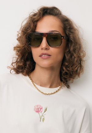 Kobieta z kręconymi włosami nosi duże okulary przeciwsłoneczne w oprawkach z masy perłowej, biały top z różowym wzorem kwiatu oraz złoty łańcuszek na neutralnym tle.