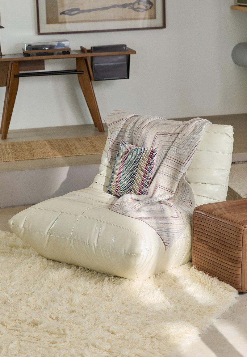 Scaun de lounge supradimensionat, cu tapițerie de culoare crem, cu un design matlasat, acoperit cu o pătură cu un model și o pernă decorativă, așezat pe un covor pufos.