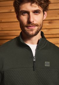 Tmavě zelený pulovr s kvartovým zipem vyrobený z texturované látky, s kontrastní panelou s geometrickým vzorem a označením značky.