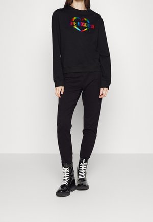Woman wearing black sweatpants and a black sweatshirt with a rainbow heart and "LOVE MOSCHINO" text, paired with black patent lace-up boots.