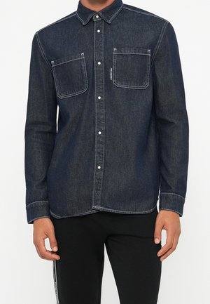 Homme portant une chemise en denim foncé à manches longues avec boutons et deux poches sur la poitrine, ainsi qu'un pantalon noir.