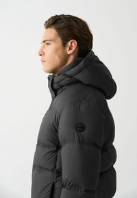 Jachetă puffer neagră cu un design texturat, matlasat, glugă și un patch rotund cu logo pe brațul stâng. Izolată pentru căldură.