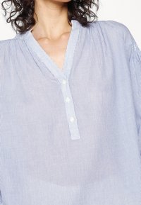 Chemise rayée bleu et blanc avec un col en V, texture douce, et boutons sur le devant. Coupe ample avec des fronces aux épaules.