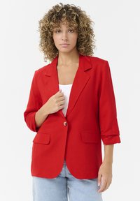 Blazer rouge en tissu léger, avec un col cranté, deux poches avant et une fermeture à un bouton. Manches retroussées.