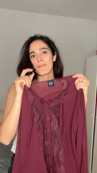 Γυναίκα κρατάει μια μπλούζα μπορντό με δαντέλα και κουμπιά, δείχνοντας την ετικέτα της μάρκας GAP σε ουδέτερο εσωτερικό χώρο.