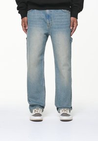 Person klädd i ljusblå raka jeans med sidofickor och vita sneakers, händerna avslappnat vid sidorna, på vit bakgrund.