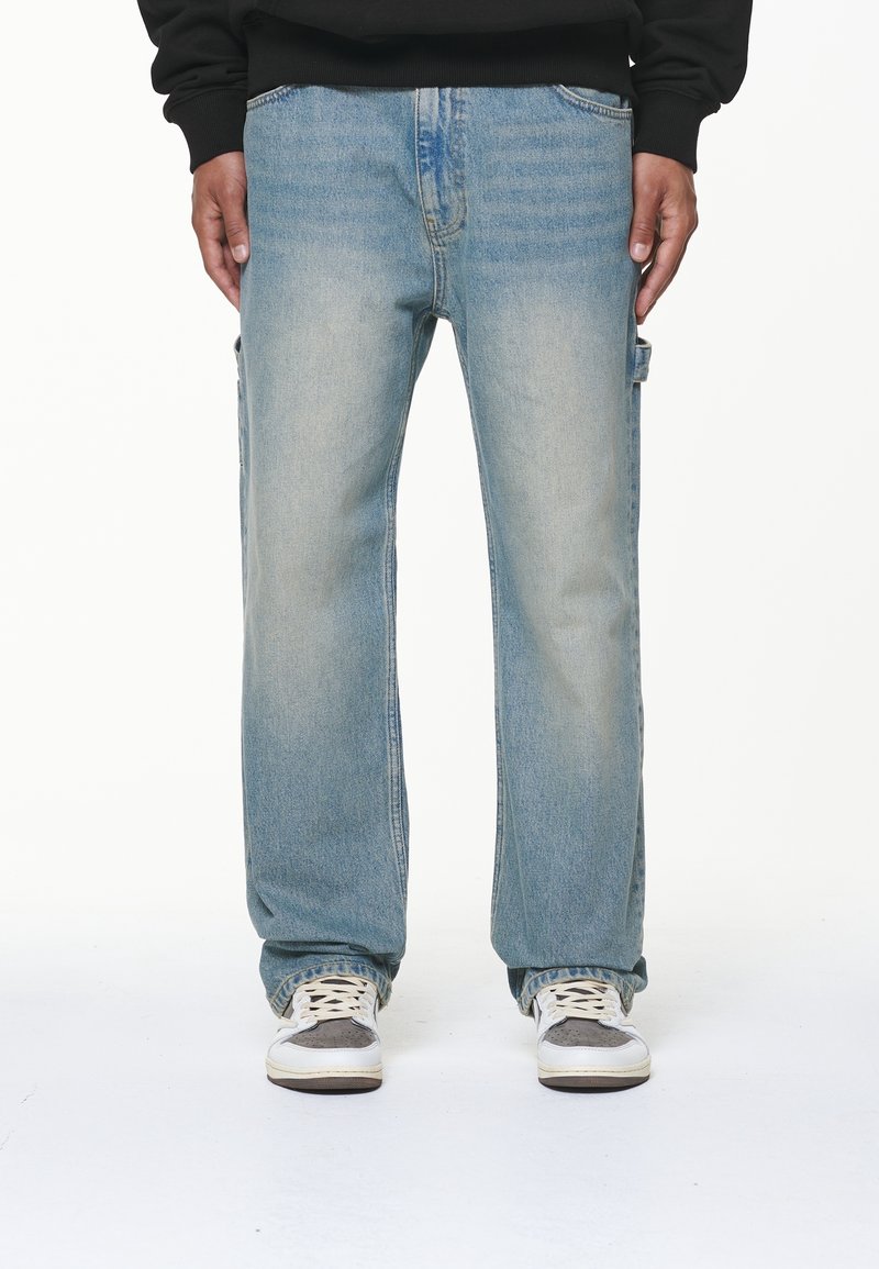 Person klädd i ljusblå raka jeans med sidofickor och vita sneakers, händerna avslappnat vid sidorna, på vit bakgrund.