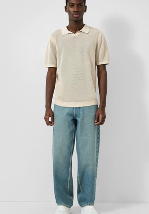 Junger Mann steht frontal und trägt ein beigefarbenes, kurzärmeliges Polo aus Strick, lockere blaue Jeans und weiße Sneaker vor einem einfarbigen Hintergrund.