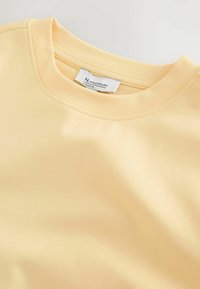 Sweatshirt en coton jaune clair avec un col rond, une texture lisse et des accents cousus. Étiquette visible à l'intérieur du col.