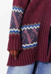 Cardigan en tricot bordeaux avec poignets côtelés et manches à motifs comprenant des accents bleus, blancs et gris dans un design géométrique.