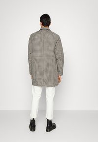 Abrigo largo a cuadros en tonos grises con corte recto y cuello, combinado con pantalones blancos y botas negras. La tela tiene un patrón texturizado.