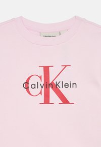 Sudadera rosa con cuello redondo que presenta una gran "K" roja y el texto "Calvin Klein" en negro. Tejido suave con una textura lisa.