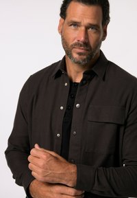 Camicia button-up marrone scuro con taschino sul petto, bottoni in metallo argentato e una consistenza morbida, indossata sopra una camicia nera.