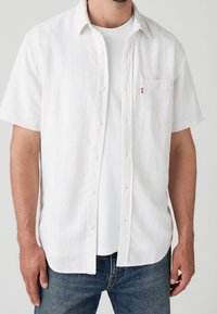 Chemise blanche à manches courtes avec des rayures verticales multicolores, boutonnée à l'avant, poche poitrine, et texture douce, portée par-dessus un t-shirt blanc uni.