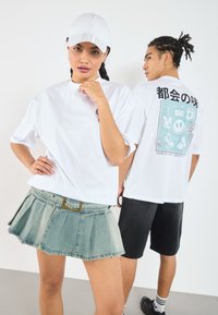 Camiseta blanca de gran tamaño con estampado gráfico, falda de mezclilla plisada azul claro y pantalones cortos negros. Los accesorios incluyen una gorra blanca con logo.