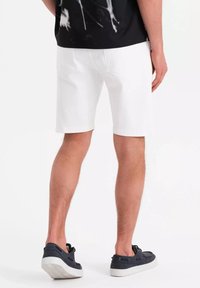 Pantalones cortos de algodón blancos con corte recto, presillas para cinturón y bolsillos laterales, combinados con zapatillas oscuras y informales. Costuras visibles que realzan el diseño.