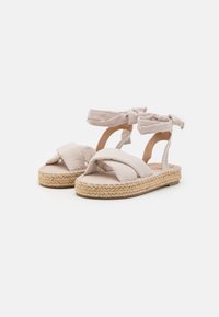 Sandalias de esparto beige con tiras de tela entrelazadas, lazos en el tobillo y una suela de yute natural. Textura suave con un perfil plano.