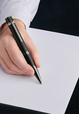 Črn svinčnik s teksturiranim držalom in kovinskimi detajli, držan nad praznim belim papirjem, ki leži na temni površini.