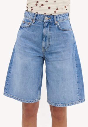 Lichtblauwe denim Bermuda-short met voor- en achterzakken, gedragen door een persoon met een lichte huidtint en een polkadot-top die is ingestopt.