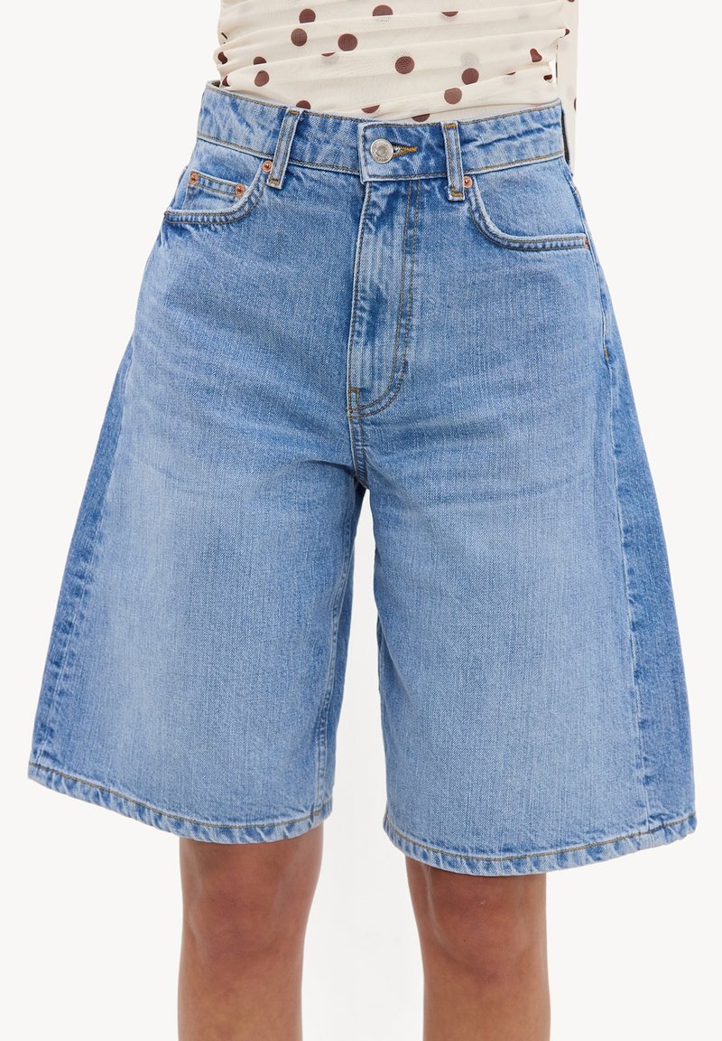 Short bermuda en jean bleu clair avec poches devant et derrière, porté par une personne à la peau claire et un haut à pois rentré.