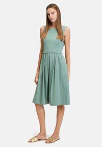 Ein ärmelloses, knielanges Kleid aus weichem grünen Stoff mit einem taillierten Oberteil und einem plissierten Rock, kombiniert mit goldenen Ballerinas.