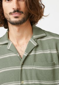 Chemise rayée verte avec un tissu texturé, présentant des rayures horizontales blanches, un col et une poche poitrine. Deux boutons de fermeture visibles.