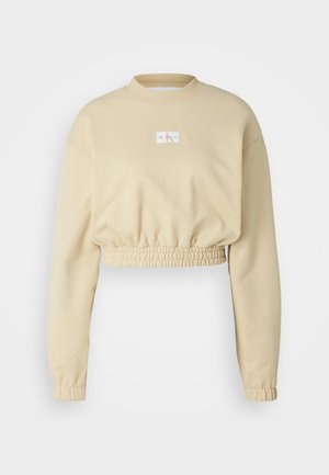 Cropped beige sweatshirt with long sleeves, gathered hem, and a small logo patch on the chest. Made from soft, textured fabric.