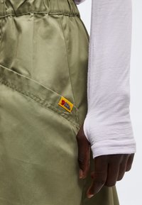 Main posée à côté d'un pantalon vert olive avec une étiquette logo Fjällräven jaune et rouge, portant une chemise blanche à manches longues avec des trous pour les pouces.