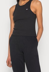 Svart ribbad linne med rundad halsringning. Har en liten broderad logotyp på bröstet. Materialet ser ut att vara stretchigt och figurnära.