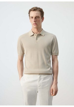 Un homme se tient face à nous, portant un polo beige rayé à manches courtes et un pantalon blanc sur un fond clair uni.