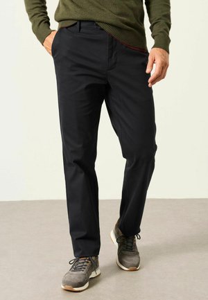 Mann trägt schwarze Hose, grünen Pullover und graue Sneakers, steht mit einer Hand in der Tasche auf einem schlichten Boden.