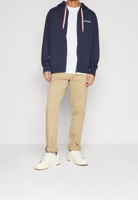 Felpa con zip di colore navy, dotata di tasche frontali e lacci rossi, abbinata a pantaloni beige e sneakers bianche. Design semplice e casual.
