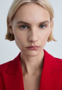 Červený blazer s ostrými klopami, strukturovaným designem a hladkým materiálem. Modelka má minimální make-up, krátké blond vlasy a světlou pleť.