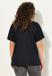 Top nero dalla vestibilità comoda con maniche a balze, realizzato in tessuto morbido. Sul retro presenta un design semplice, abbinato a leggings grigi con motivo.